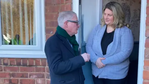 Mayor Richard Parker, wearing a coat, scarf and glasses, stands talking to Holly Farmer, in a black top and light blue jacket. They are in the doorway of her home in Friar Park. There is a window on one side with blinds visible and the garden outside is reflected in the glass.