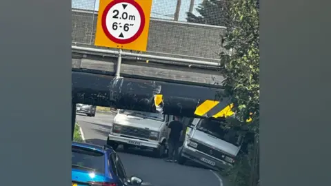 James Fuller A white van stuck under the Stonea Road Bridge with another one at its side, falling into the verge. A man is standing with his back to the viewer between the vans. The bridge has a sign above it by saying it is 2m or 6ft6in tall