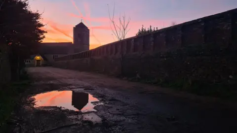 A wall leads through the shot to a small church which stands in front of a sunset sky full of purple and orange hues. There are contrails from aircraft criss-crossing the scene. In the foreground a puddle on the path catches a reflection of the top of the church tower and the sunset behind it.