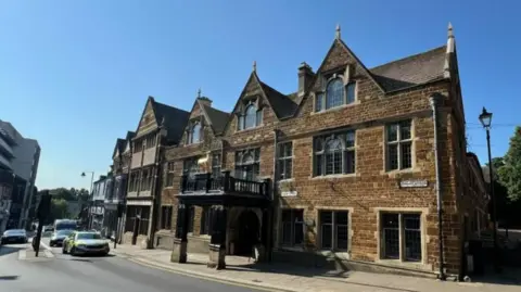 Steve Hubbard/BBC The frontage of the Hind Hotel in Wellingborough - a brown, listed brick building with a wooden canopy outside the main entrance that goes over the pavement. Cars queue at traffic lights on the road outside.