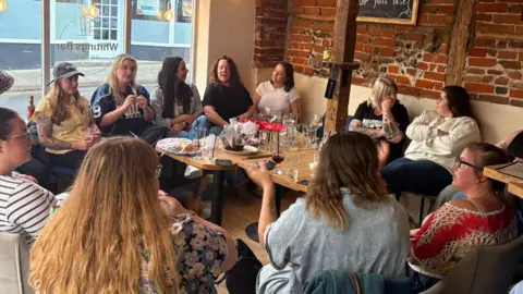 Lauren Emms A group of women sit in a large circle in a pub talking to each other. 