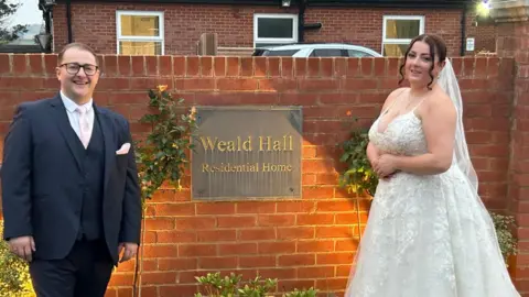 Contributed Jack and Lauren are pictured outside the care home in front of its entrance sign in their wedding attire. The care home entrance sign sits on a red brick wall and is written in gold writing against a square black backdrop.