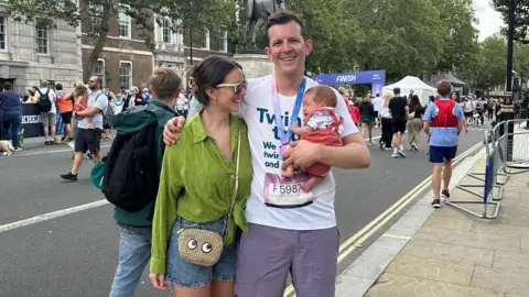 Twins Trust Mr and Mrs Quinton Smith, with Mr Quinton Smith holding baby Finn whole wearing running clothes and a medal, at the finish line of a running race