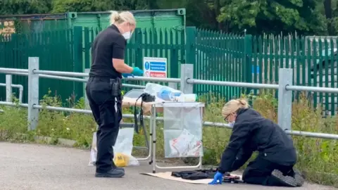 Two female police officers wearing surgical masks examine items next to a camping table holding plastic evidence bags
