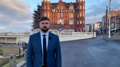 Chris Webb, with short, dark brown hair and a beard, wears a blue jacket, light blue shirt and dark blue tie. He is photographed standing on the promenade in front of the Metropole Hotel in Blackpool.