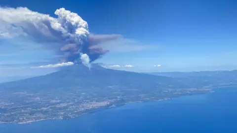 Jon Meek A picture of Mount Etna erupting from a plane. There is grey and black cloud coming out of the top of the volcano. The sky is blue. The ground is green and grey. The sea is a darker blue than the sky.