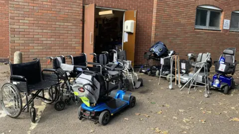 A range of wheelchairs, scooters and walkers outside the red brick Harborough Shopmobility