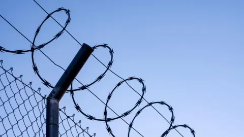 Getty Images Razor-wire on top of a tall metal fence.