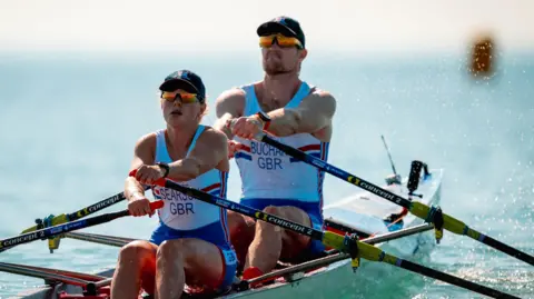 Benedict Tufnell Two athletes in a rowing boat on open water, each holding oars. They are wearing matching blue and white uniforms with “GBR” printed on them, indicating representation for Great Britain. The water is calm and the sky is bright.