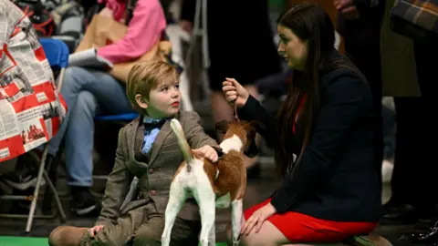 Getty Images A young boy in a tweed suit sitting on the floor with his mum who is wearing a red dress and black blazer. A small, white and brown dog is stood between the pair. They are sitting on a green carpet.