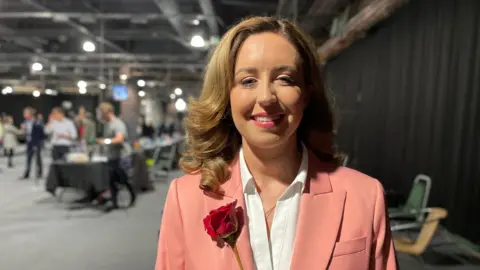 BBC/Mark Ansell Sally Jameson stands in a hall. She wears a salmon-coloured blazer and white blouse with a rose fixed to her right side. She has curly, shoulder-length brown hair.