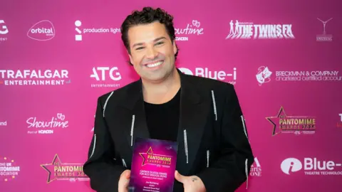 Ian Olsson Joe McElderry is holding his award and smiling brightly at the camera. He has short, curly black hair and is wearing a black blazer with silver tassels on in. The award is an A5 pink square which reads: "The Pantomime Awards 2026".