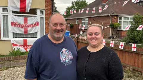 Luke Deal/BBC A man and a woman stood next to each other in front of a home that is decorated in England flags and bunting. They are smiling and looking into the camera. 