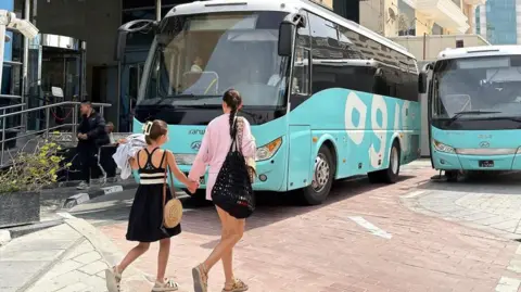 A woman and a girl walk in front of buses prepared for stranded airline passengers outside a hotel in Doha, Qatar. Photo: 1 March 2026