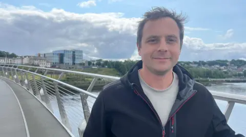 a man stands on a bridge wearing a blue jacket. a river is behind him, and a number of buildings can be seen in the background