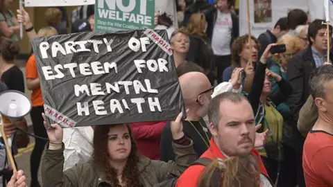 Getty Images People demonstrating in the street in Manchester, England. One campaigner is holding up a placard that says "PARITY OF ESTEEM FOR MENTAL HEALTH".