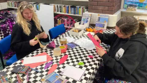 Two women, each wearing black tops sitting either side of a table with a black and white checked tablecloth. They're cutting feather shapes out of pink and purple fabric and placing them on the table. 