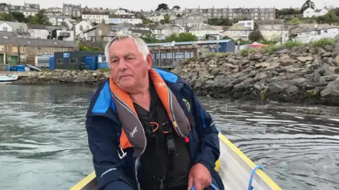 A man wearing a lifejacket coxes a wooden gig boat . He holds the ropes to steer the vessel.