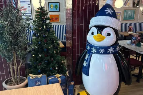 Prezzo A penguin decoration wearing a blue hat and scarf standing near a Christmas tree inside a restaurant.