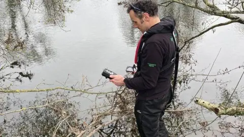 Environment Agency In the picture, a person is standing at the edge of a calm river, holding a handheld device that looks like water‑testing equipment. The person is wearing outdoor waterproof clothing, including dark trousers and a dark jacket with logos on the sleeves and back. 