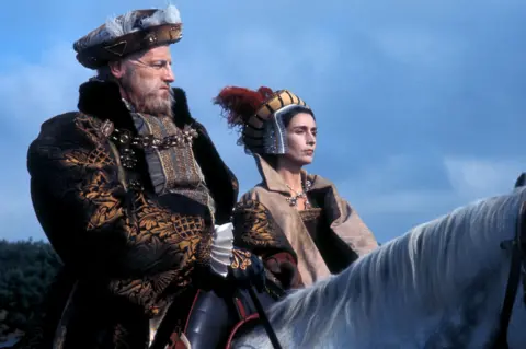 A still from a TV drama showing a male actor dressed as King Henry VIII, in an elaborate black and amber jacket and feathered cap, riding a white horse. Alongside him is a female actor dressed as Queen Catherine Parr, wearing a suede cloak over a black dress, and a structured headdress made of white, gold and black cloth, topped by red feathers.