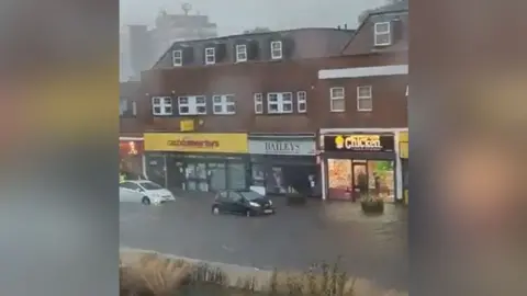 Rudd1979/X Cars driving on Dunstable High Street, which has been covered in floodwater due to heavy rain. There are shops in the background.