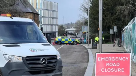 A van and a red emergency road closure sign on a road. In the background are a police car and a person in a high-vis jacket.