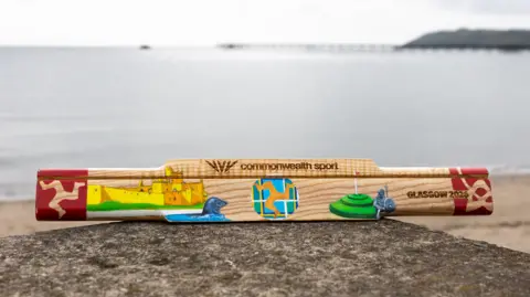 Vannin Photos The wooden baton is sitting on a stone wall top, in front of a beach with the tide in on a very calm and grey day. In the distance you can see an iron pier to the right. The baton has different artistic pictures on it in different colours - they include Peel Castle, a seal, and the Manx triskelian symbol. 