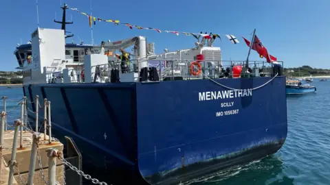 A large blue vessel branded Menawethan Scilly is seen in the sea with a blue sky behind it, flags across its deck and a harbour seen to the left 