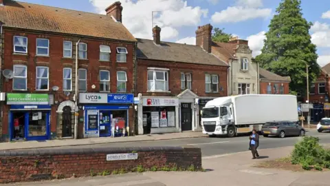 Google A single-carriageway urban road with a line of shops on the ground floor and two and three-storey terraced buildings. There is a white lorry parked (the wrong way round) outside a shop. There are cars moving past it, and a person in a blue sweater and black coat is walking past and looking at a phone. FORD END ROAD is written on a white sign on a low brick wall in the foreground.