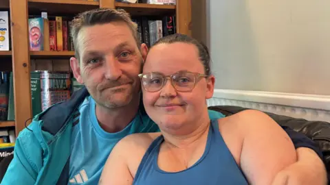Mark and Elen take a selfie on a sofa with a bookcase full of books behind them. Elen has her hair tied back and is wearing glasses and a blue vest top, and Mark has short hair and stubble and is wearing a blue jacket and blue Adidas t-shirt. 