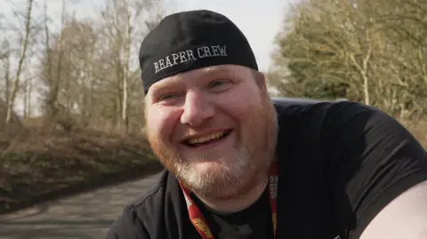 Shaun Whitmore/BBC A man smiling while wearing a black tshirt, a basbeball cap on back to front with Reaper Crew in white lettering across it