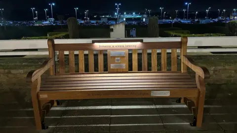 Kris Laskowski A brown wooden bench, situated in front of a low brick wall. The photo has been taken in darkness, with several street lights visible in the distance. The bench has a light panel on the top which has illuminated some black writing which says "someone is always listening". There are also several plaques installed on the bench.