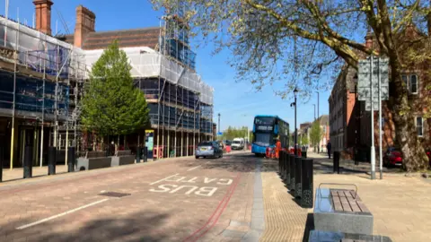 A stretch of pink coloured paved road, with the words bus gate on it, and a bus parked up in the distance. There are buildings to the left and right of the road