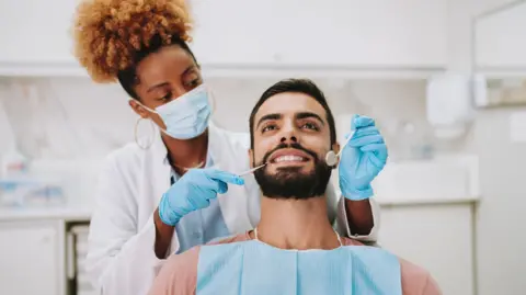A dentist, wearing a facemask, white coat and blue gloves is checking a man's teeth.