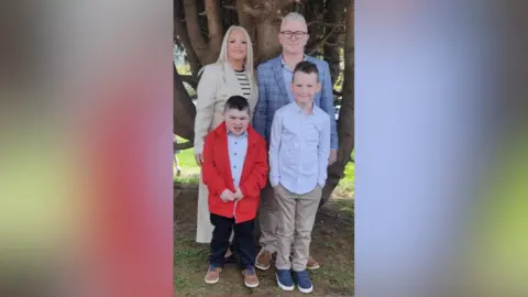 Family handout Alfie (bottom left) with his family. A woman behind him has blonde hair, wearing a beige coat and black and white striped top. A boy beside him is wearing a a light blue shirt, beige chinos and blue trainers. Alfie is wearing a red coat, light blue shirt, black trousers and brown shoes. His dad Jamie is wearing black glasses, a blue check blazer and blue shirt. They are all standing in front of a tree.