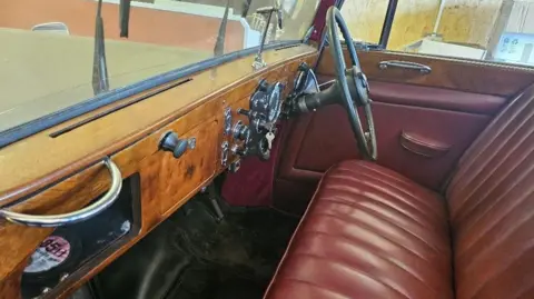 Humbert & Ellis The interior of a vintage Daimler vehicle, with red upholstery and a wooden dashboard. There is a tax disc in the lower left corner of the image.