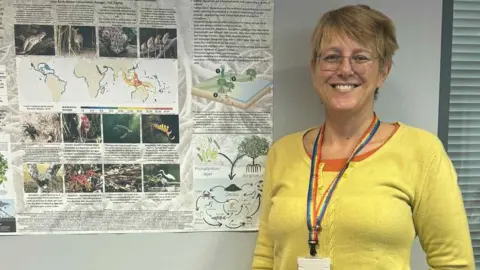 Lisa Holdsworth Lisa Holdsworth has short blonde hair and wears glasses, a yellow cardigan and a rainbow-coloured ID pass. She stands by a biology poster which is stuck on the wall. 