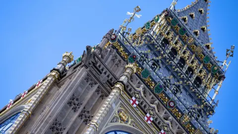 The top part of the tower with intricate gold details and green shields. There is a row of small St George's flags running above the clock face.  