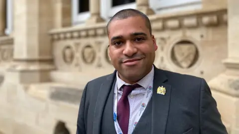 Laura Coffey/BBC A man in a black blazer, white shirt and red tie stands in front of a stone building. 