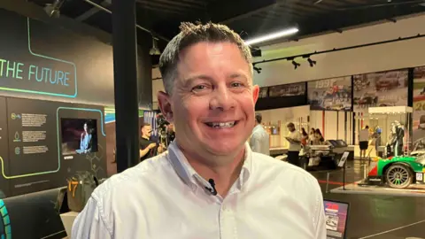 Kate Bradbrook/BBC Rob Jaina with short blond hair smiling at the camera and wearing a white shirt. There is a display behind him called "The Future" and part of a green car is visible to his left. There are people looking at exhibits behind him.