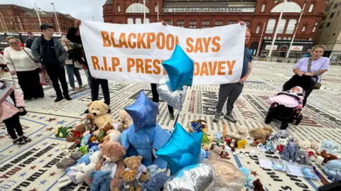 Yunus Mulla/BBC A group of people hold a white banner with the words 'Blackpool says R.I.P Preston Davey' in orange lettering. On the ground are teddy bears, blue star shaped balloons and cards. 