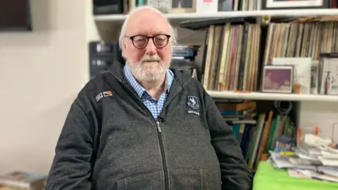 BBC Martin Shipton is wearing a light wash black zip hoody and a blue chequered shirt underneath. He has a beard and is wearing glasses. Behind him are books and portraits.
