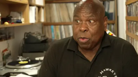 Roy Forbes-Allen in a Hawkeye Records polo shirt sits among shelves of vinyl records and turntables