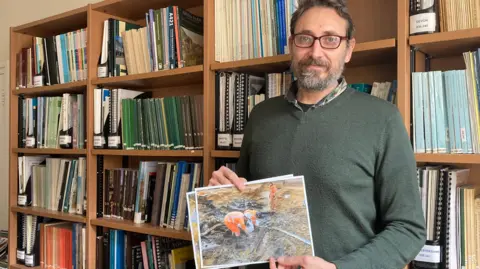 A man with a light grey beard and glasses holds photographs of an archaeological site in front of him as he stands with his back to a large bookcase