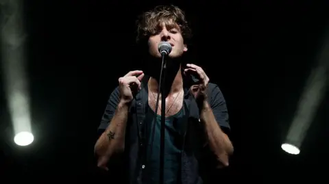 Getty Images Scottish singer Paolo Nutini singing into a microphone. He is wearing a black vest and shirt. A star tattoo is visible on his left arm