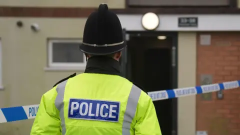 Police officer with his back turned to the camera wearing a yellow high viz police jacket and a black police helmet. He is stood in front of a property that has police tape outside it.