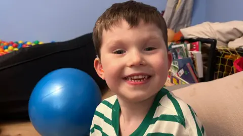 a smiling boy with toys and books behind him. He is wearing hearing aids in both ears