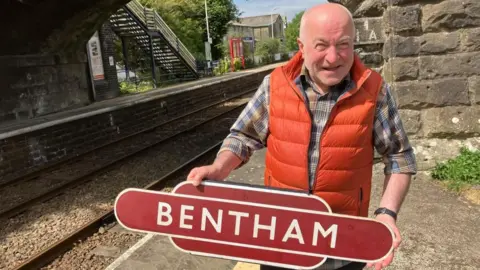 A man in a green checked shirt and orange gilet stands on a railway station platform holding a station sign which says 'Bentham' on it. In the background is the station platform.