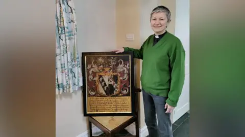 The vicar of St Leonard's, Reverend Jo Burke, stands with the returned wooden panel. It is positioned on a chair. Reverend Burke wears jeans and a green jumper over her dog collar.